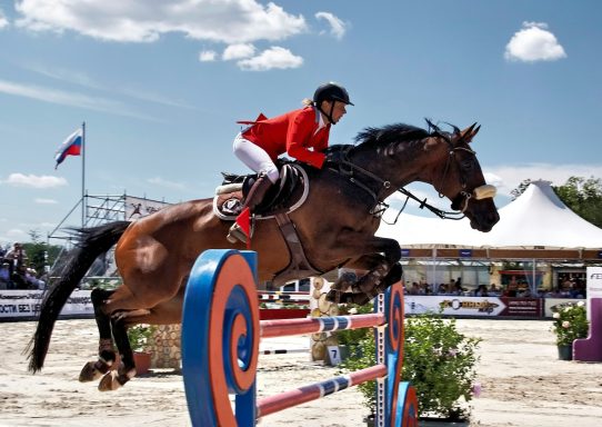 Horse and rider in red uniform jumping over a large obstacle at an equestrian event.